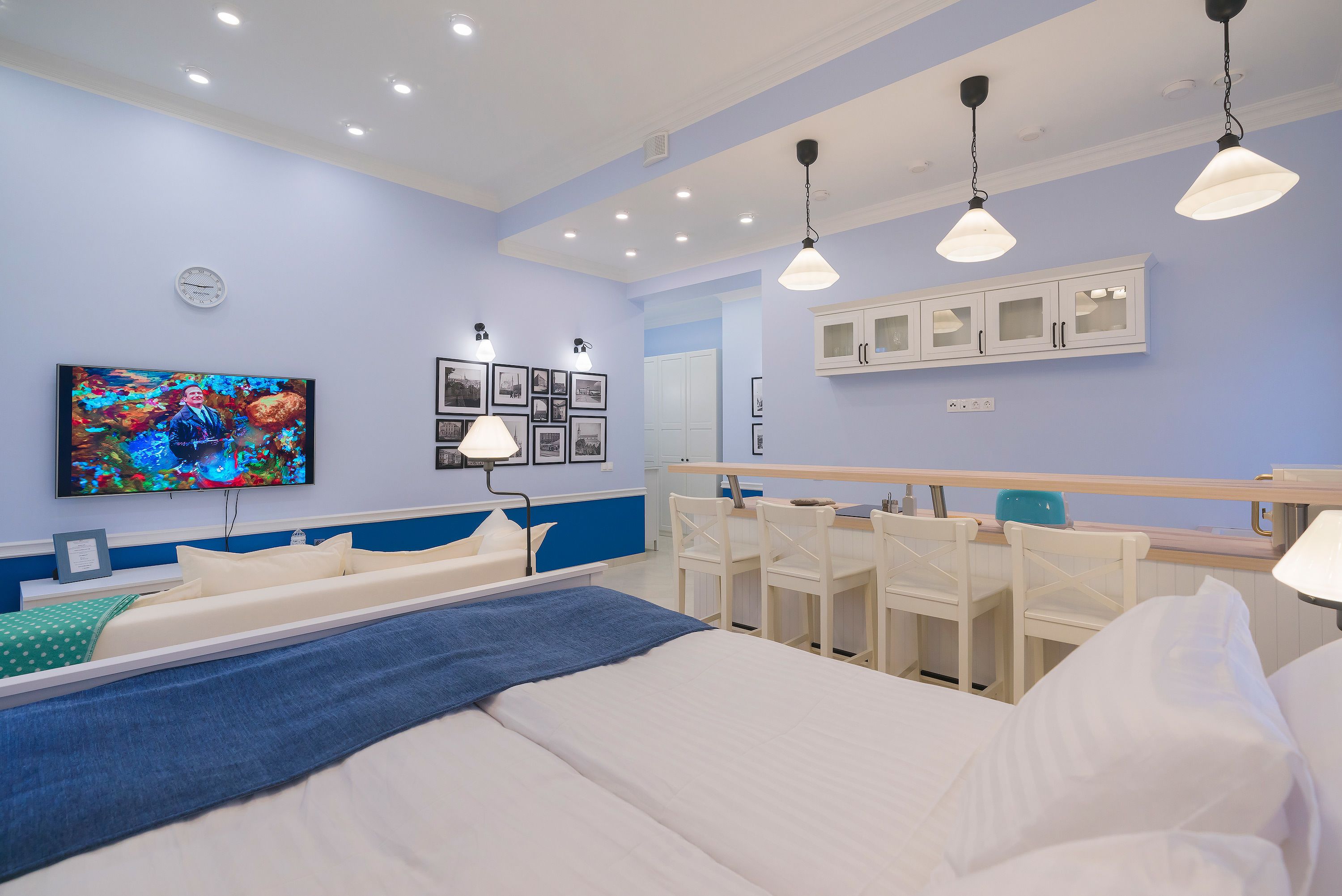 A modern hotel room with a large bed, bedside tables, a desk, and a chair. There is a television mounted on the wall, a clock, and framed pictures.
