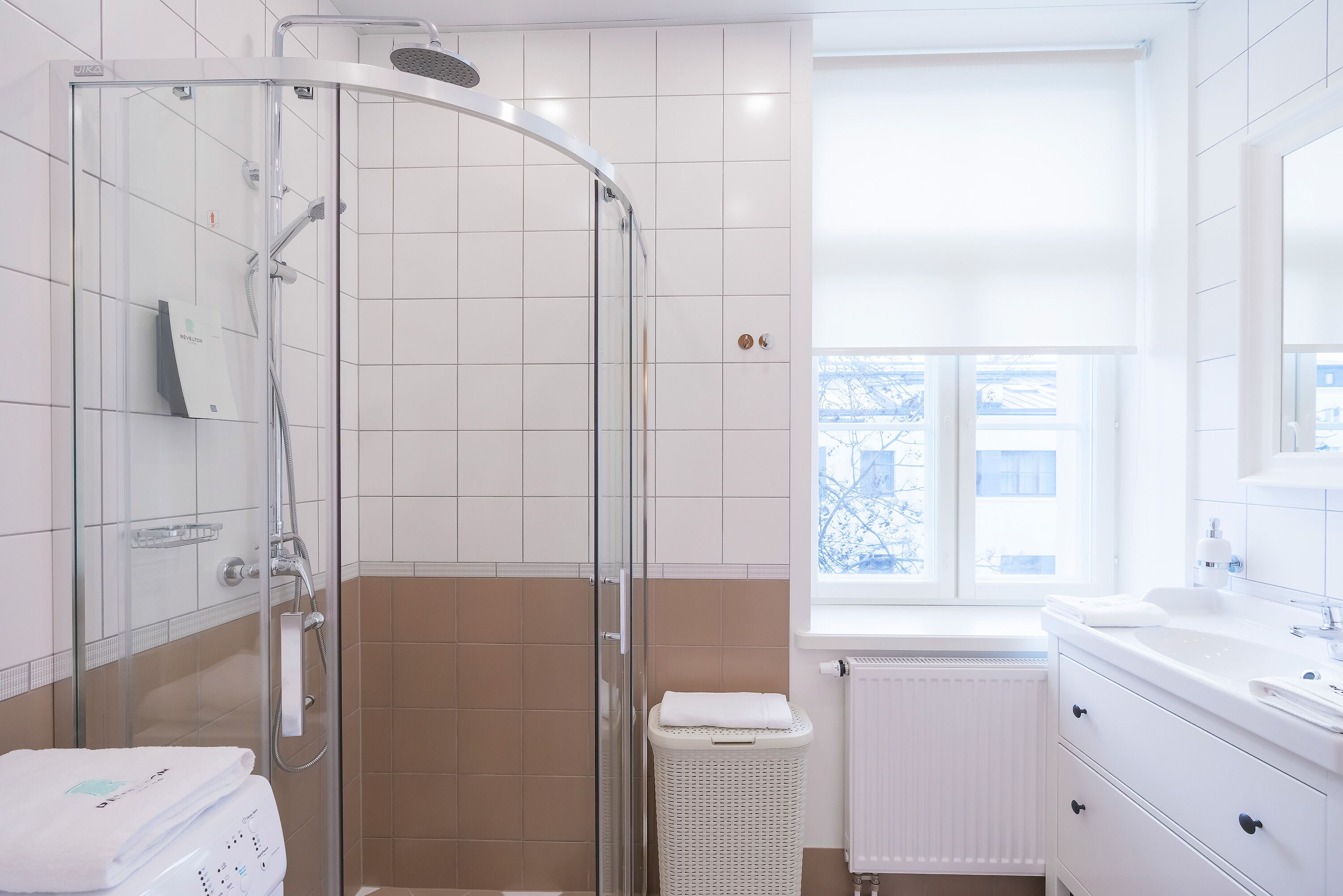 A modern bathroom with a glass-enclosed shower, white tiles, a window with a view, and a white sink.