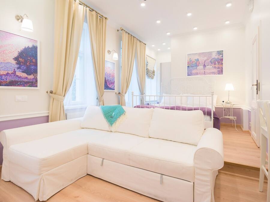 A modern, well-lit living room with a white sectional sofa, a small table with a lamp, and two large windows with curtains.