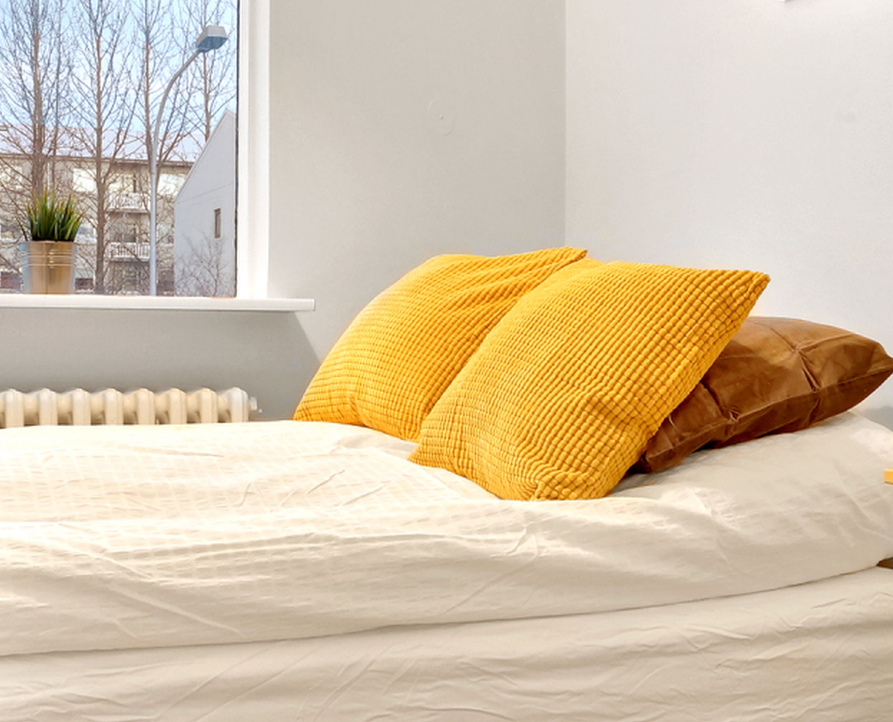 Ein Bett mit weißer Bettwäsche und zwei Kissen, eines gelb und eines braun, in einem Zimmer mit Fenster und Heizkörper.