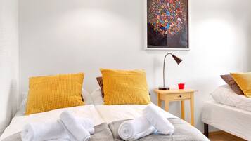 A neatly made bed with white and yellow pillows, a grey blanket, and rolled white towels. There is a small wooden nightstand with a lamp and a red cup beside the bed. A colorful abstract painting is on the wall above the bed.
