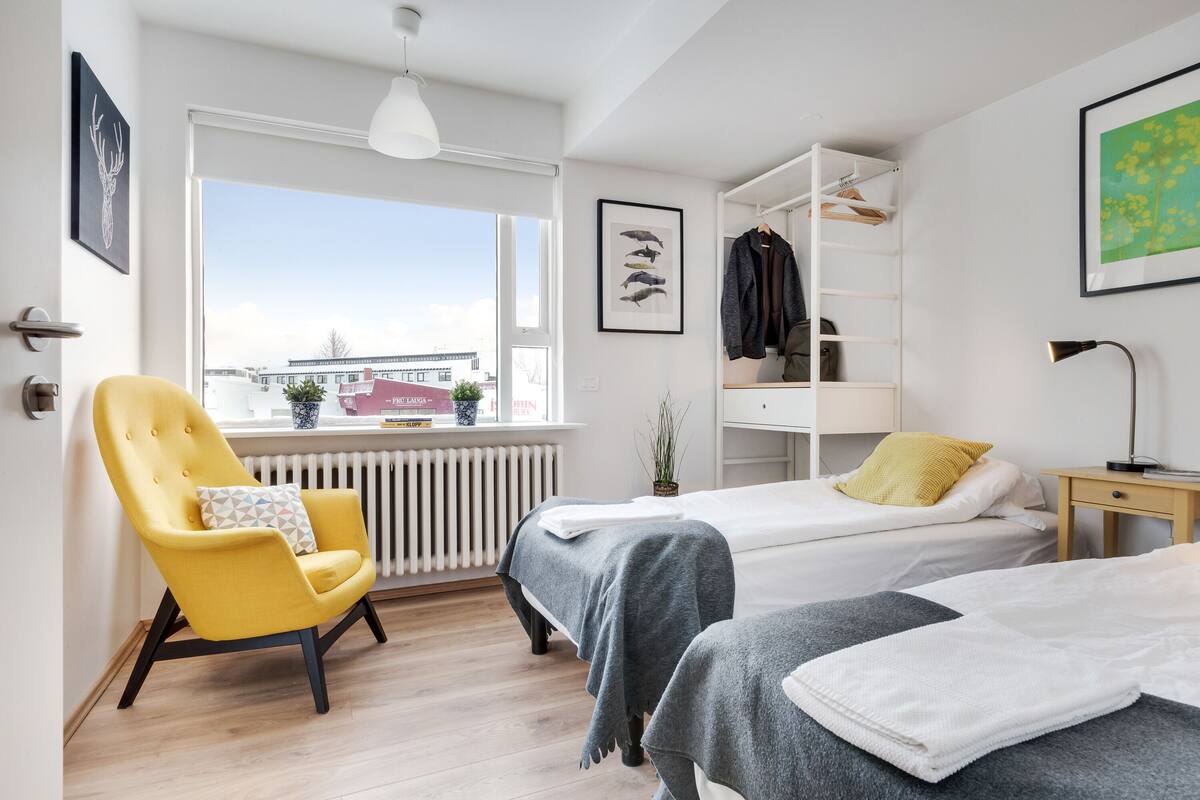 Ein modernes Hotelzimmer mit zwei Betten, einem gelben Sessel, einer Heizung und einem Fenster mit Blick auf die Stadt.