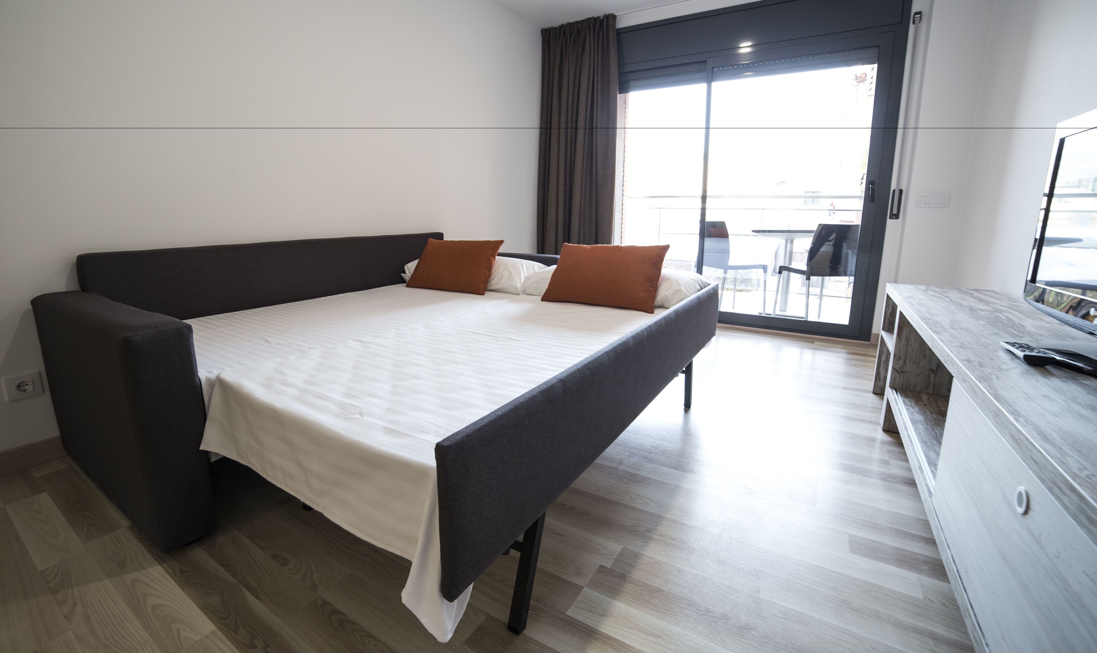 Una habitación de hotel moderna con una cama grande, un sofá oscuro, piso de madera y un balcón con mesa y sillas.