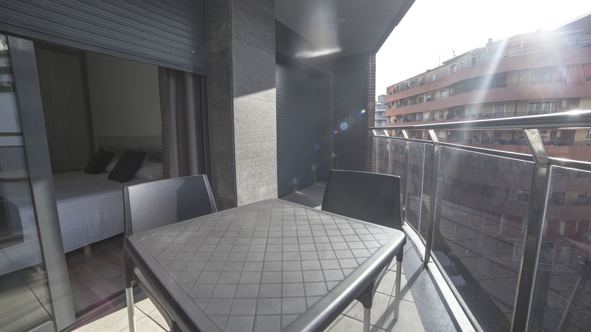 A balcony with a table and chairs, a bed with pillows, and a view of a building across the street.