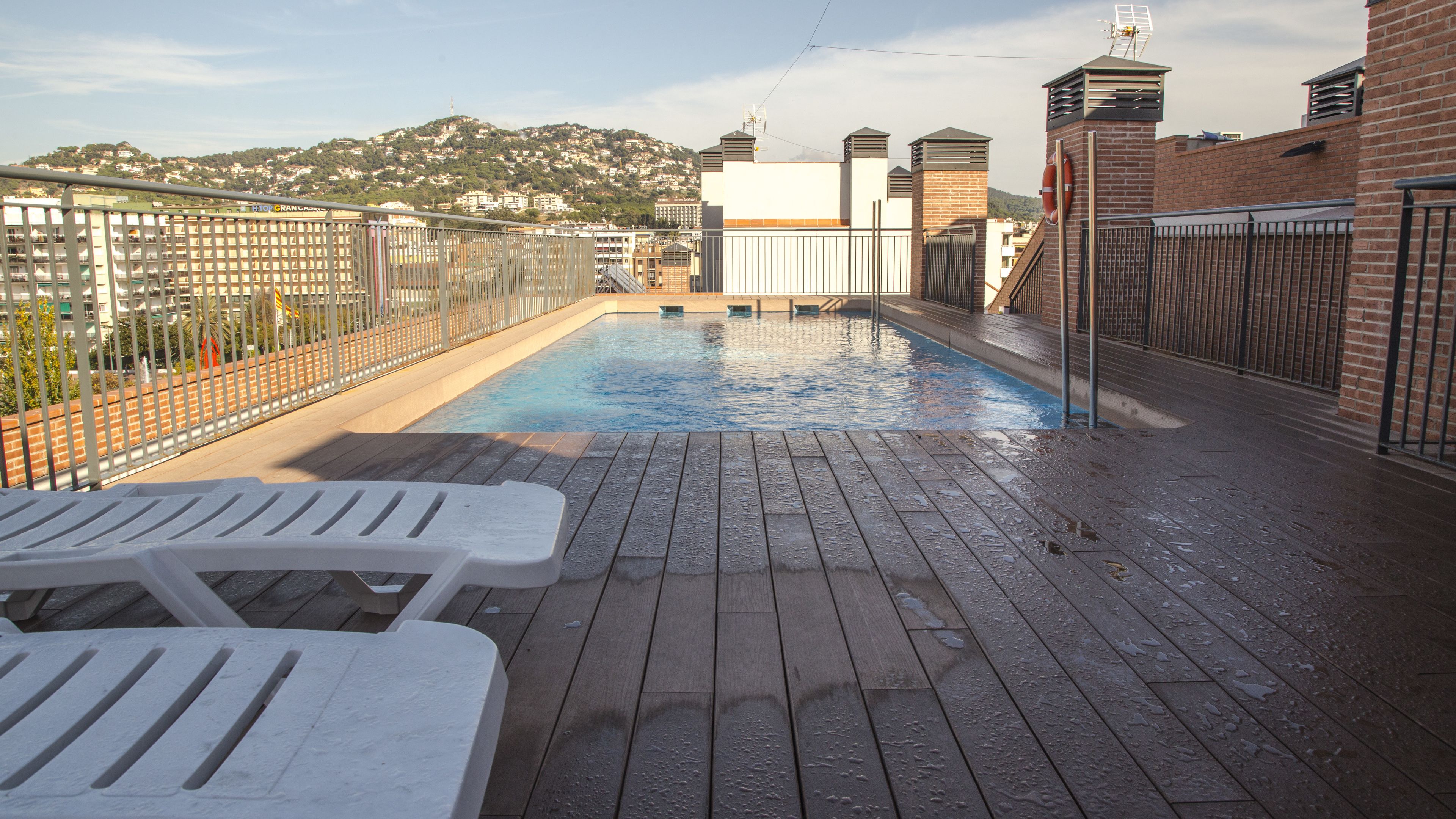 A rooftop pool with wooden deck and lounge chairs, surrounded by a metal fence and brick walls.