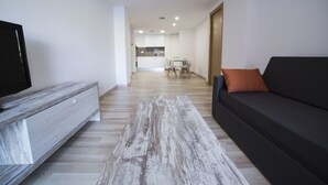 Un saló modern amb televisió de pantalla plana, un sofà gris amb un coixí taronja i una taula de centre de fusta. Es veu una cuina al fons.