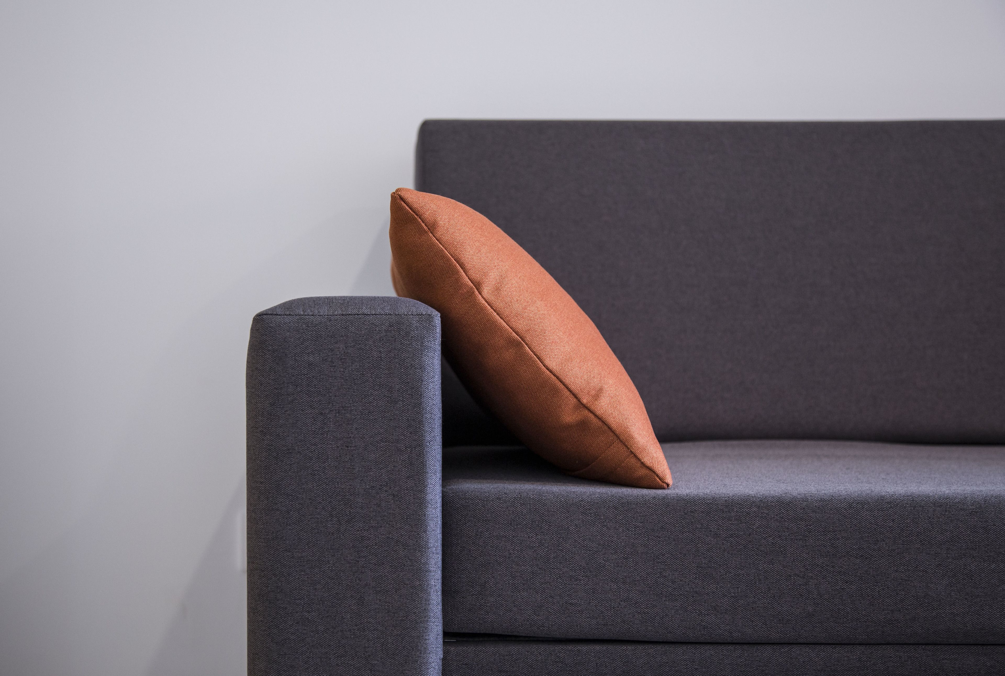 A dark grey sofa with a single brown cushion against a plain wall.