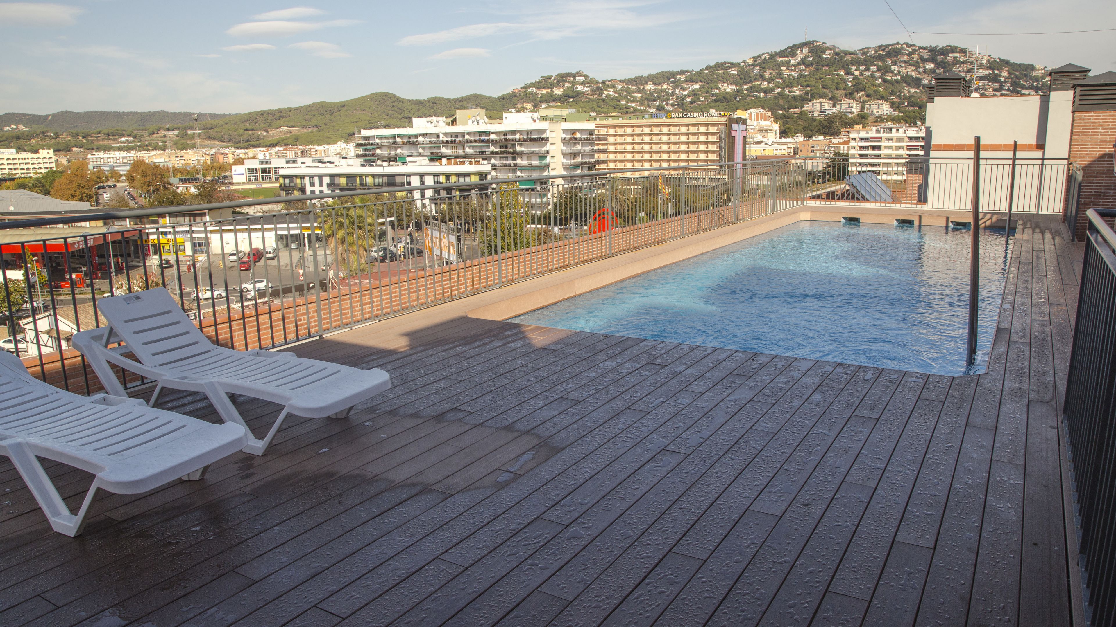 Ein Pool auf dem Dach mit zwei weißen Liegestühlen, einer Holzterrasse und Blick auf eine Stadtlandschaft mit Gebäuden und Hügeln im Hintergrund.