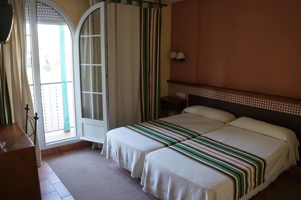 A bedroom with a double bed, a wooden headboard, a nightstand, a lamp, and a view through a window with curtains.