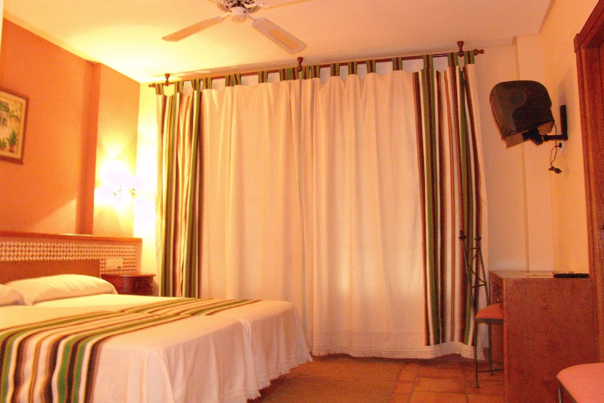 A hotel room with two beds, a ceiling fan, a desk with a chair, and a curtain.