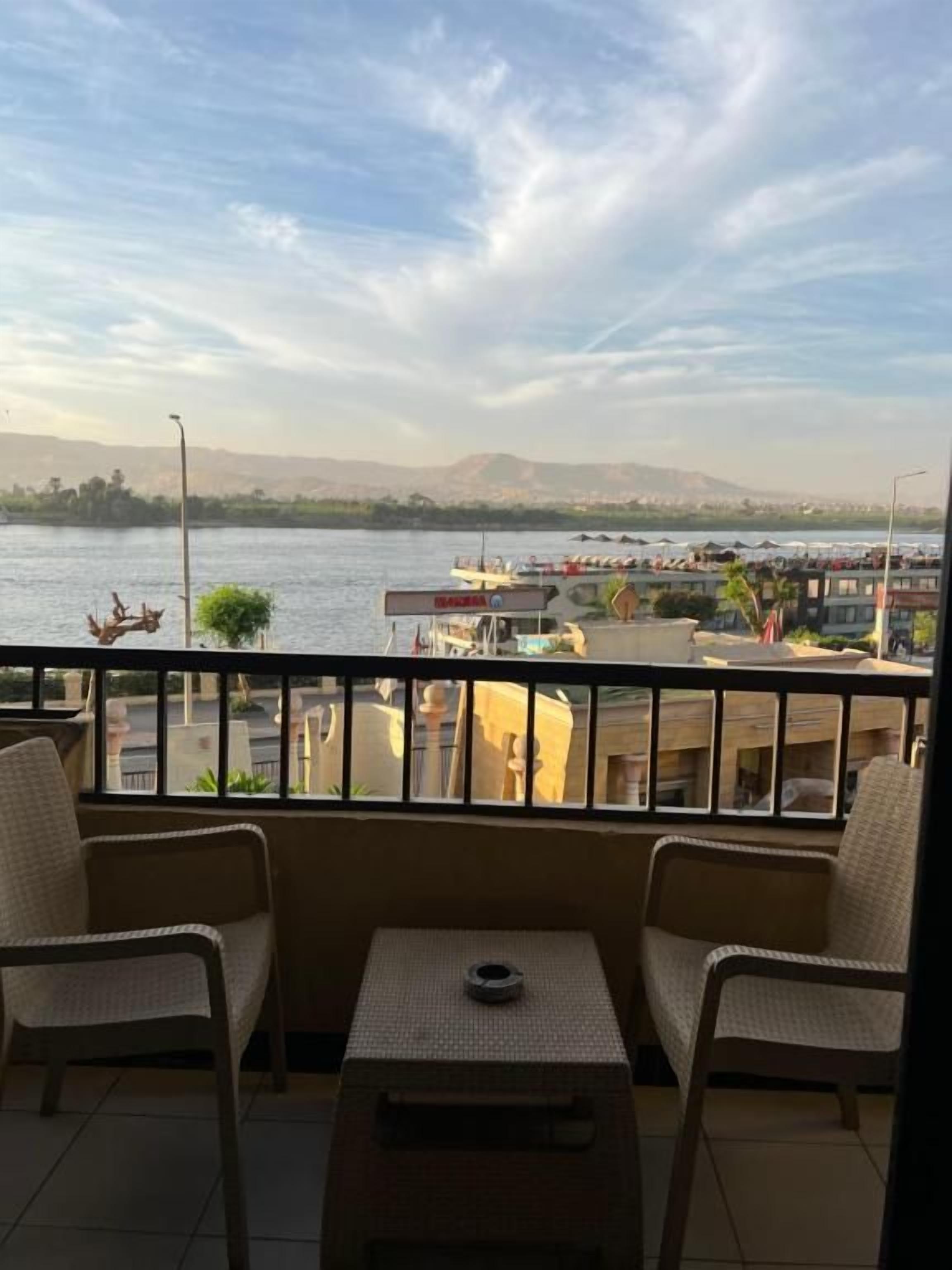 A balcony with two chairs and a small table, overlooking a body of water with boats and buildings in the distance.