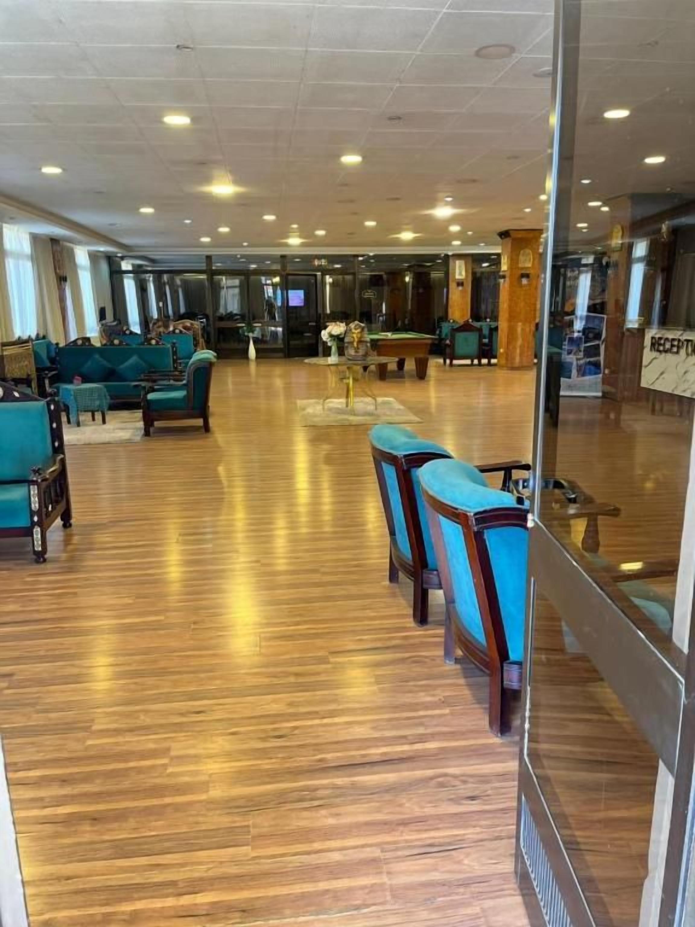 A spacious lobby with a wooden floor, seating area with blue chairs, a reception desk, and a television screen.