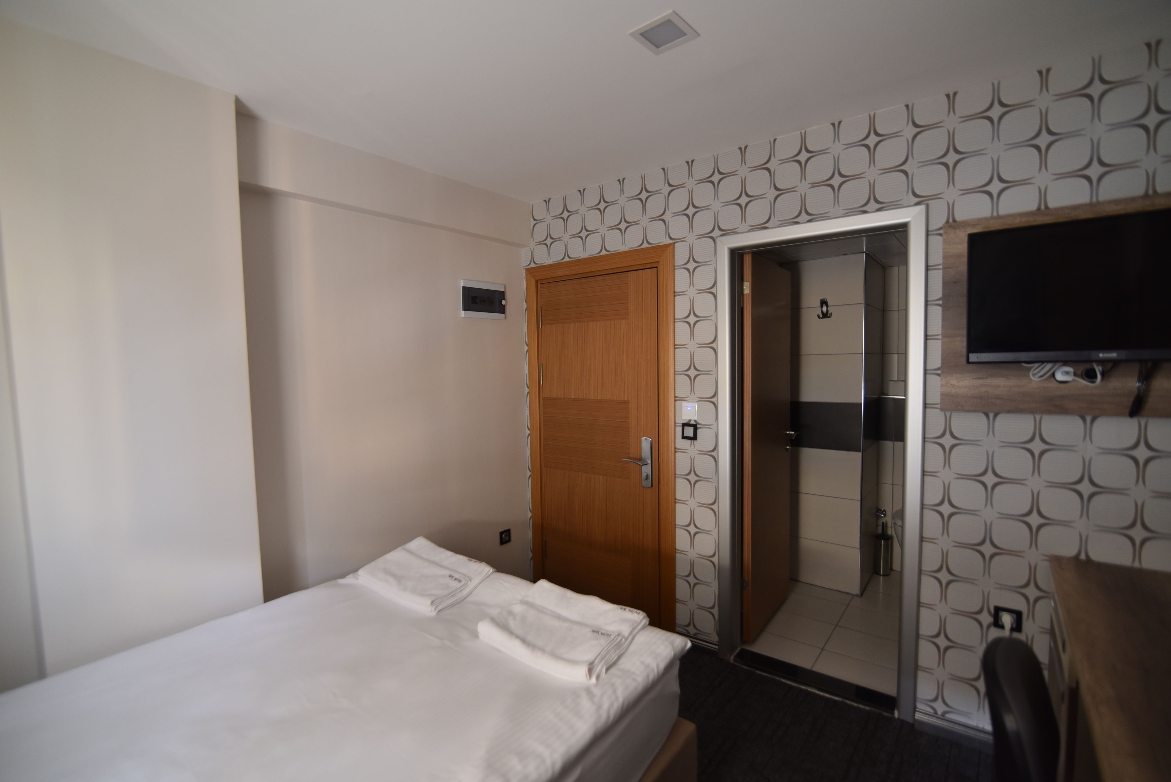 A hotel room with a bed, a wooden door, a bathroom with a mirror, and a TV mounted on the wall.