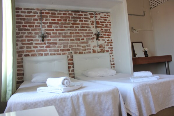 Quarto de hotel com duas camas, roupa de cama branca, parede de tijolos e mesa de cabeceira de madeira com espelho e um vaso.