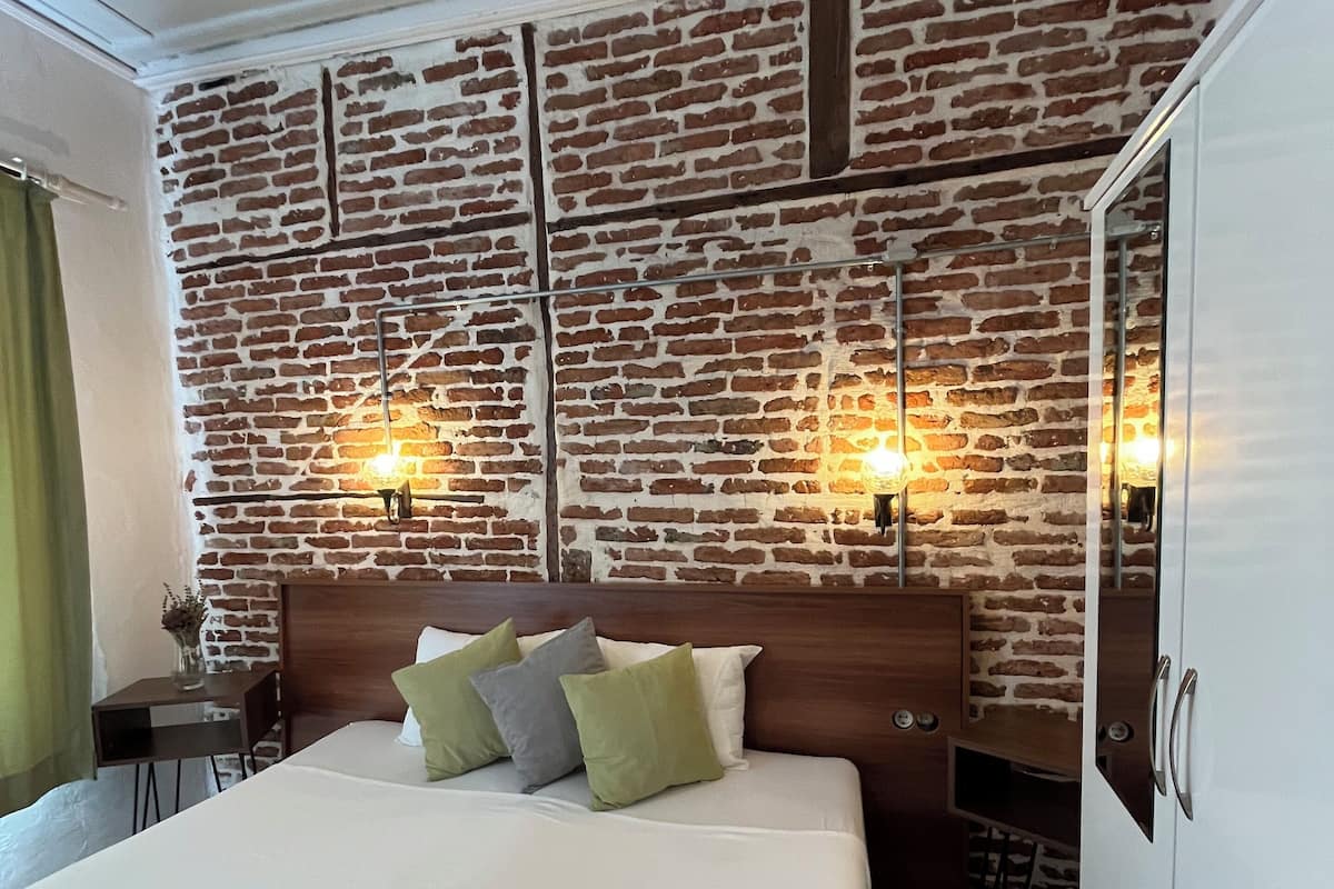 A hotel room with a brick-patterned wall, a large bed with pillows, a wooden headboard, a small table with a vase, and a wardrobe.