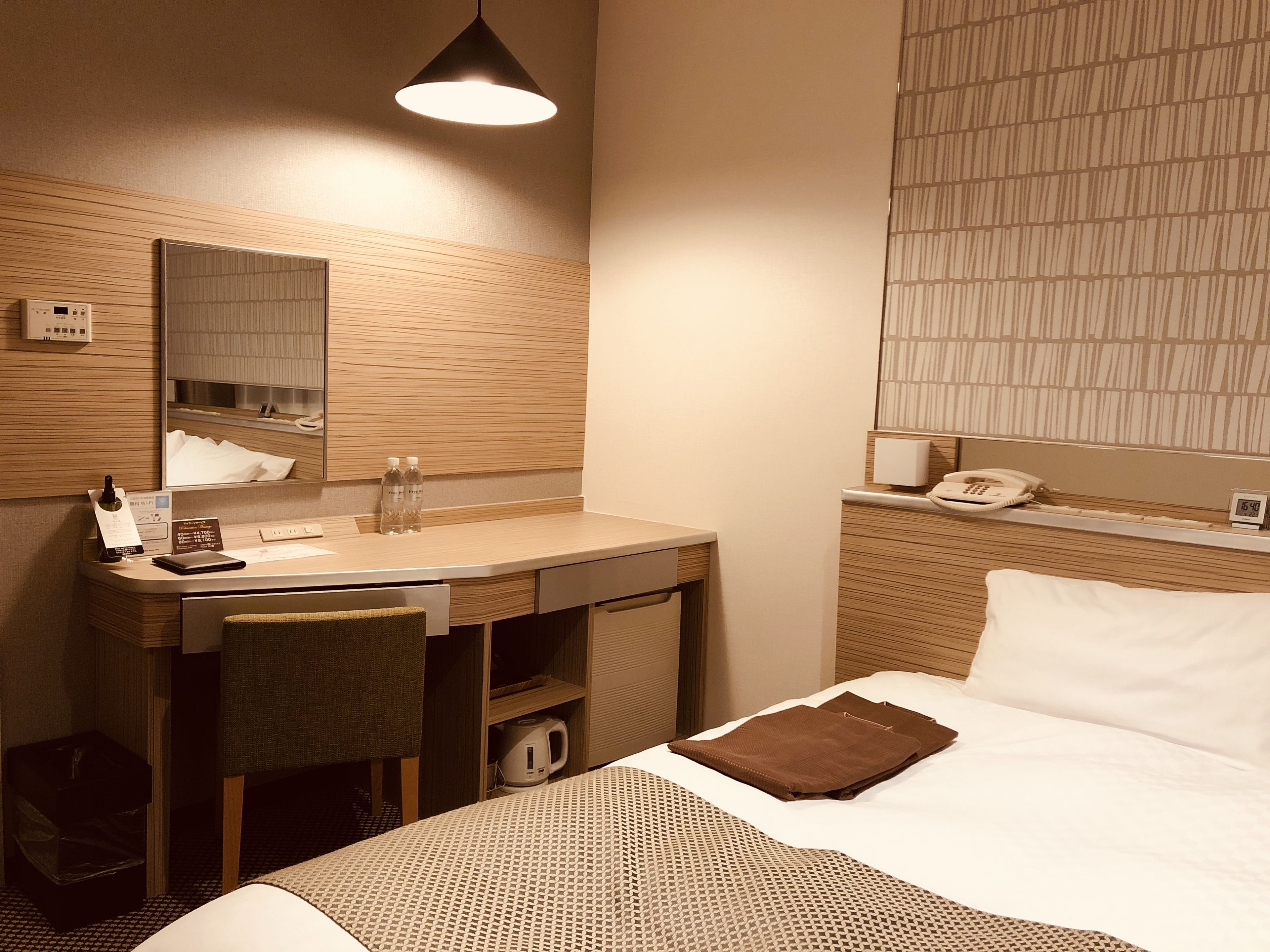 A hotel room with a wooden desk, a mirror, a telephone, and a bed with a patterned bedspread.
