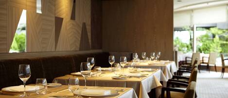 A restaurant with long tables set for dining, featuring a view of greenery outside.