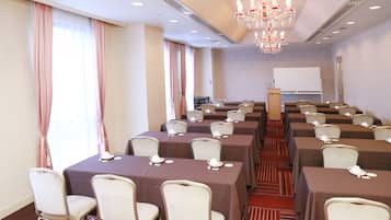 Un salón de conferencias con filas de mesas y sillas, una pantalla de proyector y una araña.