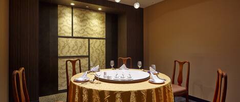 A round table with a gold tablecloth, surrounded by wooden chairs, in a room with dark wood paneling and a large window with patterned curtains.