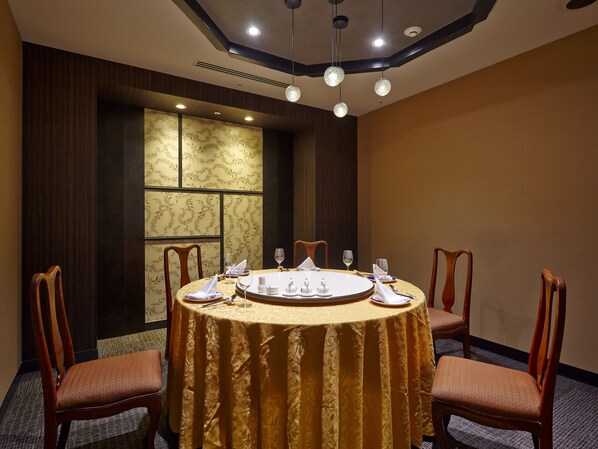 A round table with a gold tablecloth, surrounded by wooden chairs, in a room with dark wood paneling and a large window with patterned curtains. - Sendai Kokusai Hotel (Sendai)