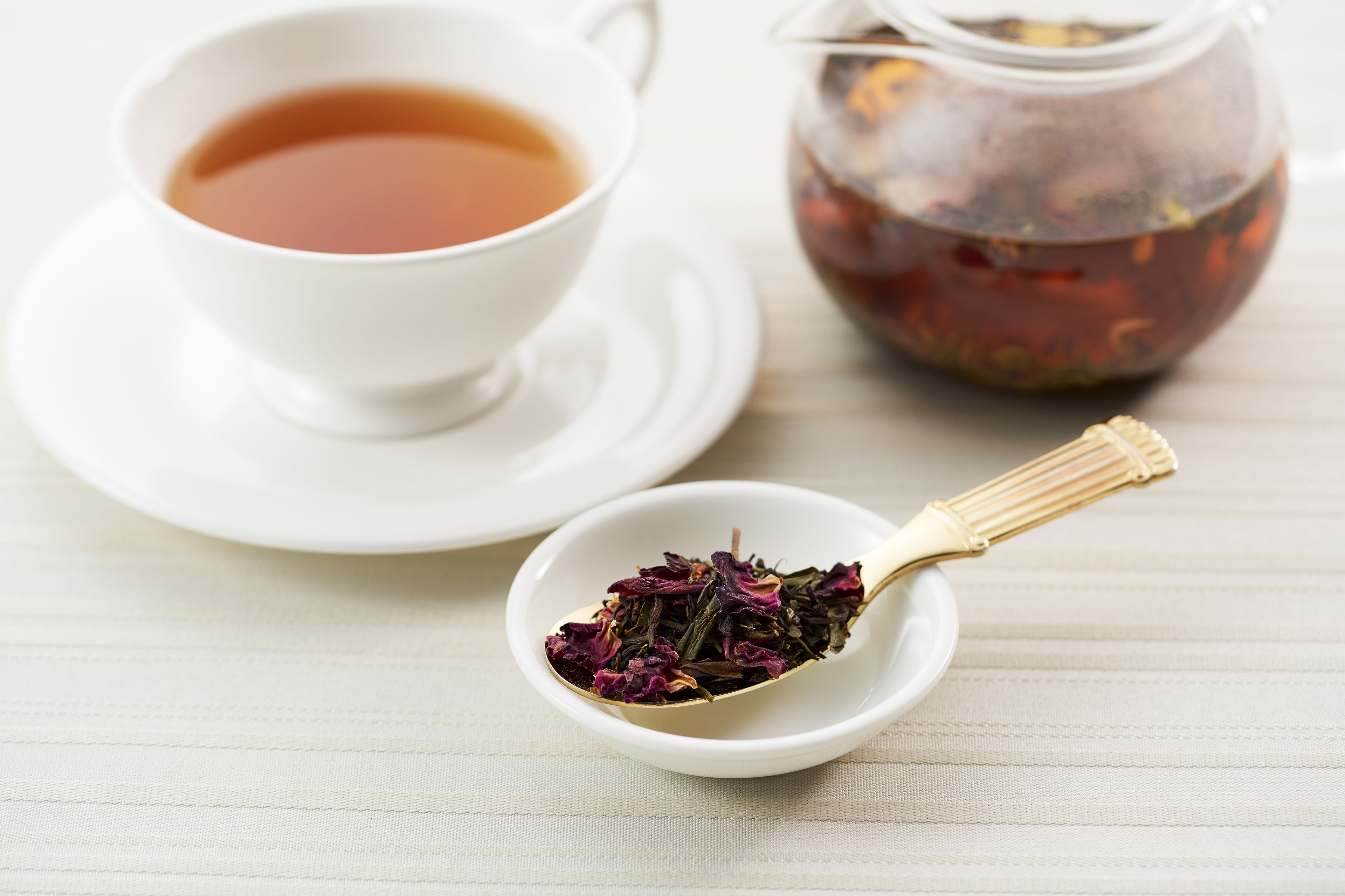 A white cup of tea on a saucer, a small white bowl with tea leaves, and a glass jar of tea on a wooden surface.