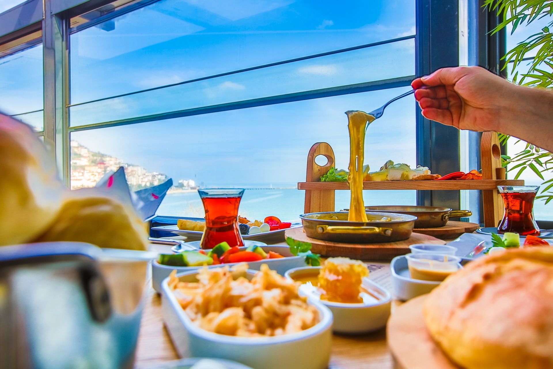 Una mesa con diversos platos y bebidas, una mano sosteniendo una cuchara y vista al mar a través de una ventana.