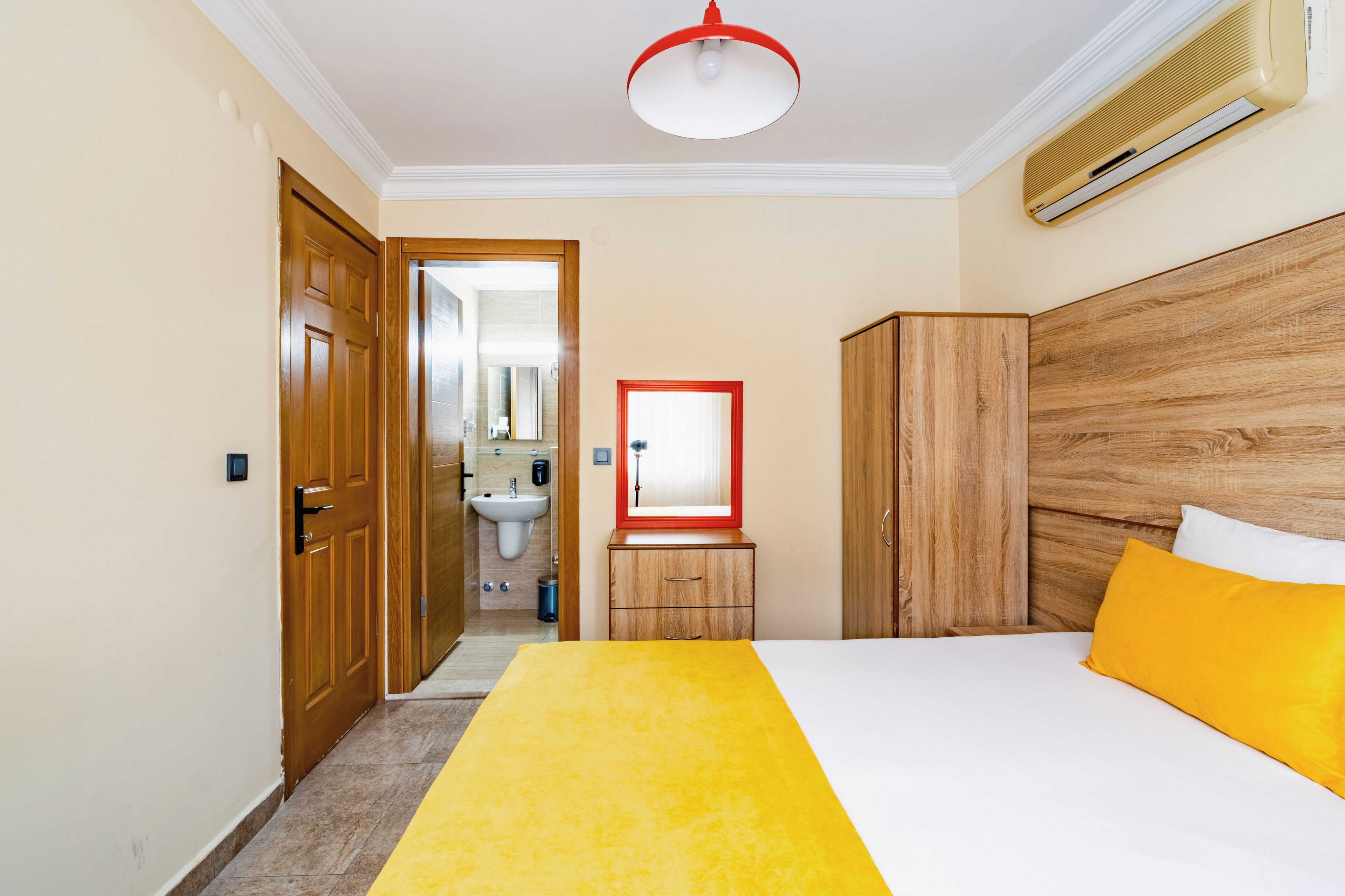 A hotel room with a wooden wardrobe, a bed with white and yellow bedding, a red-framed mirror, and a ceiling light.