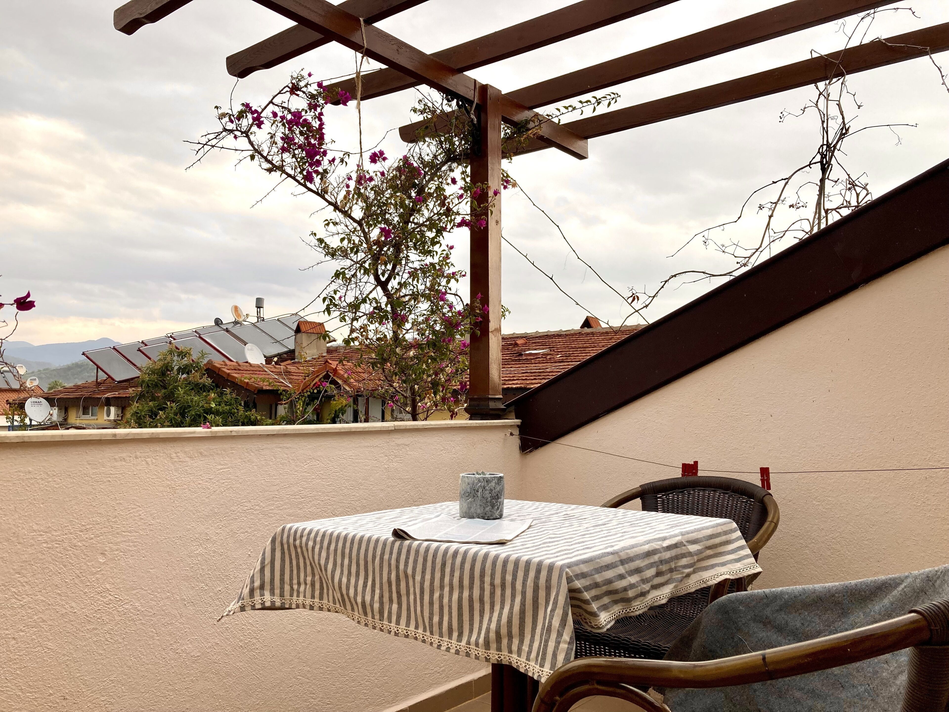 A rooftop terrace with a table and chairs, a pergola, and a view of houses and mountains in the background.