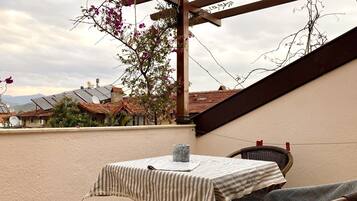 A rooftop terrace with a table and chairs, a pergola, and a view of houses and mountains in the background.