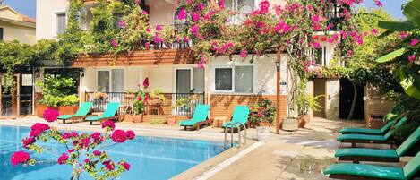 Un espace aménagé au bord de la piscine, avec des chaises longues et une piscine, le tout entouré d’un bâtiment doté de balcons ornés de plantes en fleurs.