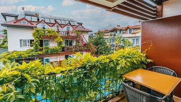 A balcony with a wooden table and wicker chairs, overlooking a swimming pool and neighboring houses.