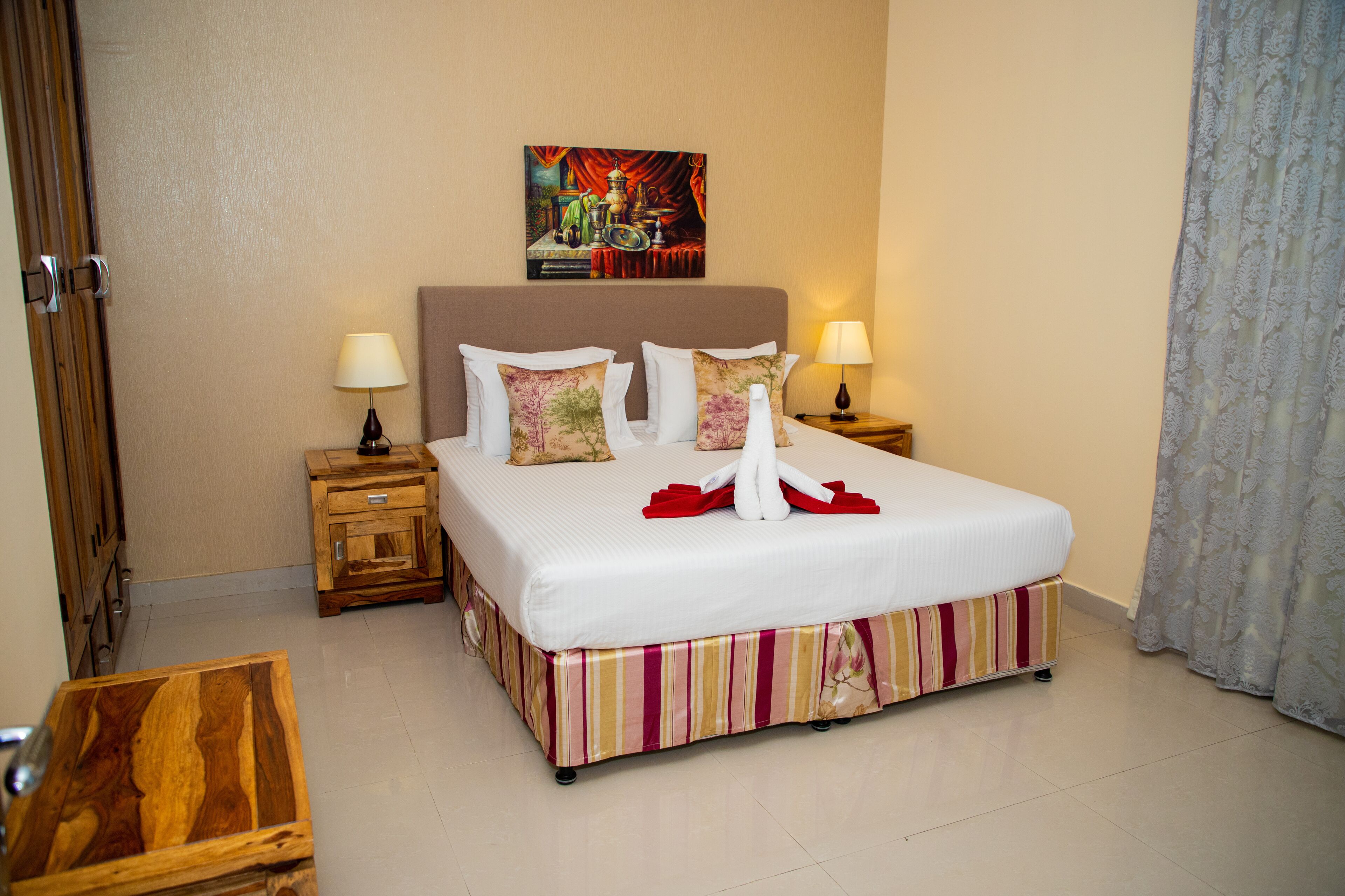 A neatly made bed with a striped bed skirt, two bedside tables with lamps, a wooden nightstand, and a painting on the wall.