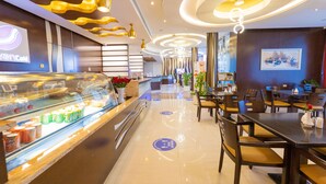 Un moderno vestíbulo de hotel con un mostrador de recepción central, una vitrina con productos alimenticios y un área de descanso con mesas y sillas.