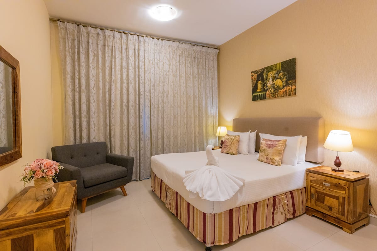 A hotel room with a bed, a chair, a nightstand, a lamp, a painting on the wall, and a curtain.