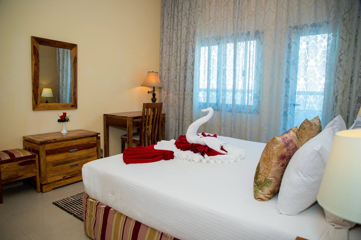 A hotel room with a neatly made bed, a wooden nightstand with a lamp, a chair, and a mirror on the wall.