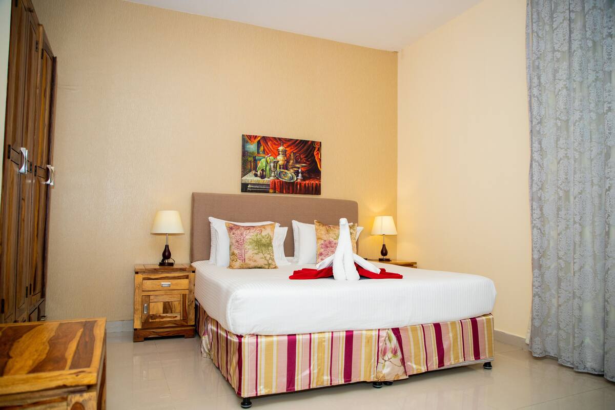 A hotel room with a large bed, two bedside tables with lamps, a wooden wardrobe, and a painting on the wall.