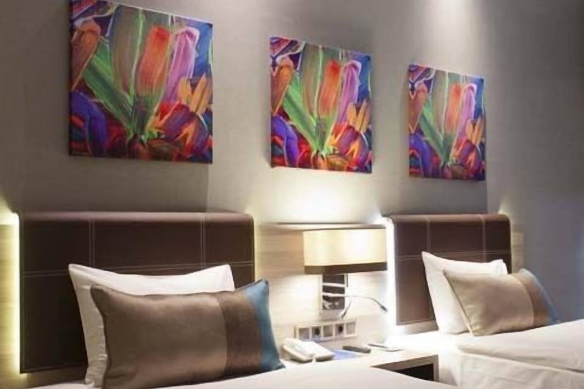 A hotel room with two beds, each with a headboard, a bedside table, and a lamp. There are three colorful abstract paintings on the wall above the beds.