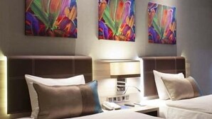 Habitación de hotel con dos camas, cada una con cabecera, mesita de noche y lámpara. En la pared sobre las camas hay tres cuadros abstractos y coloridos.