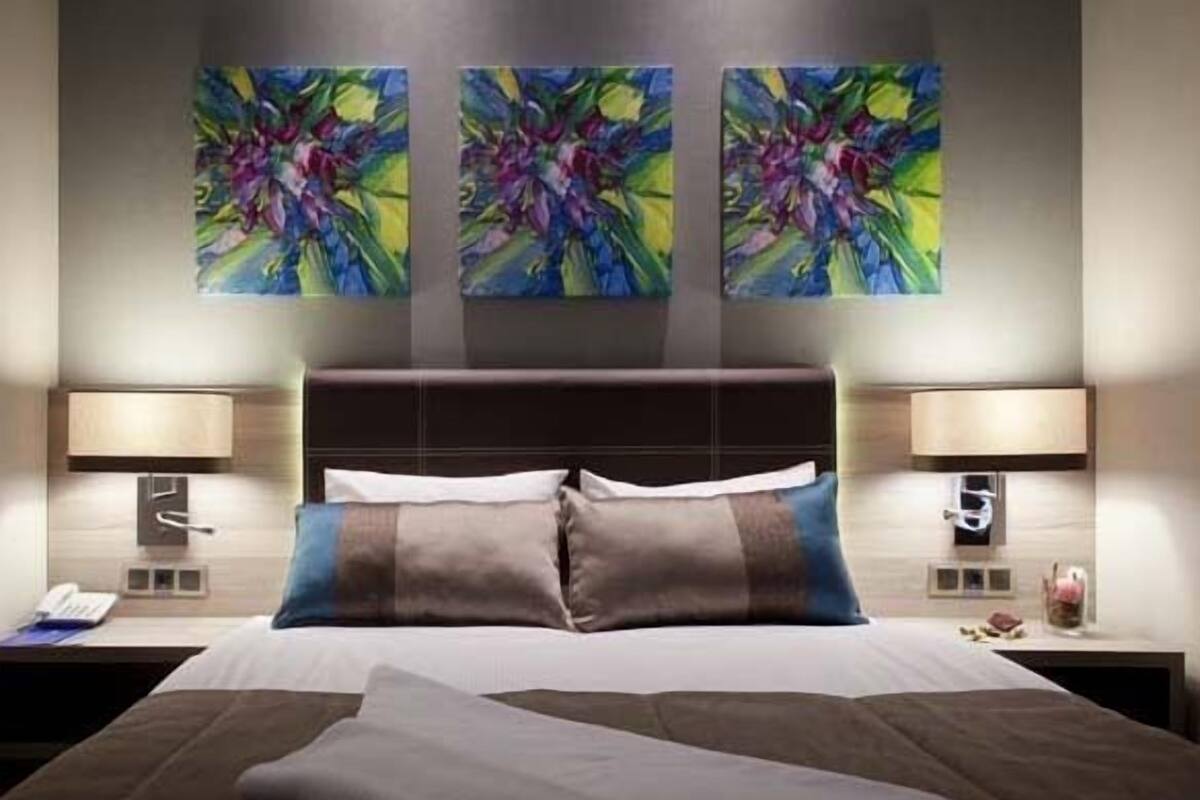 A hotel room with a large bed, two bedside tables, and three abstract paintings on the wall.