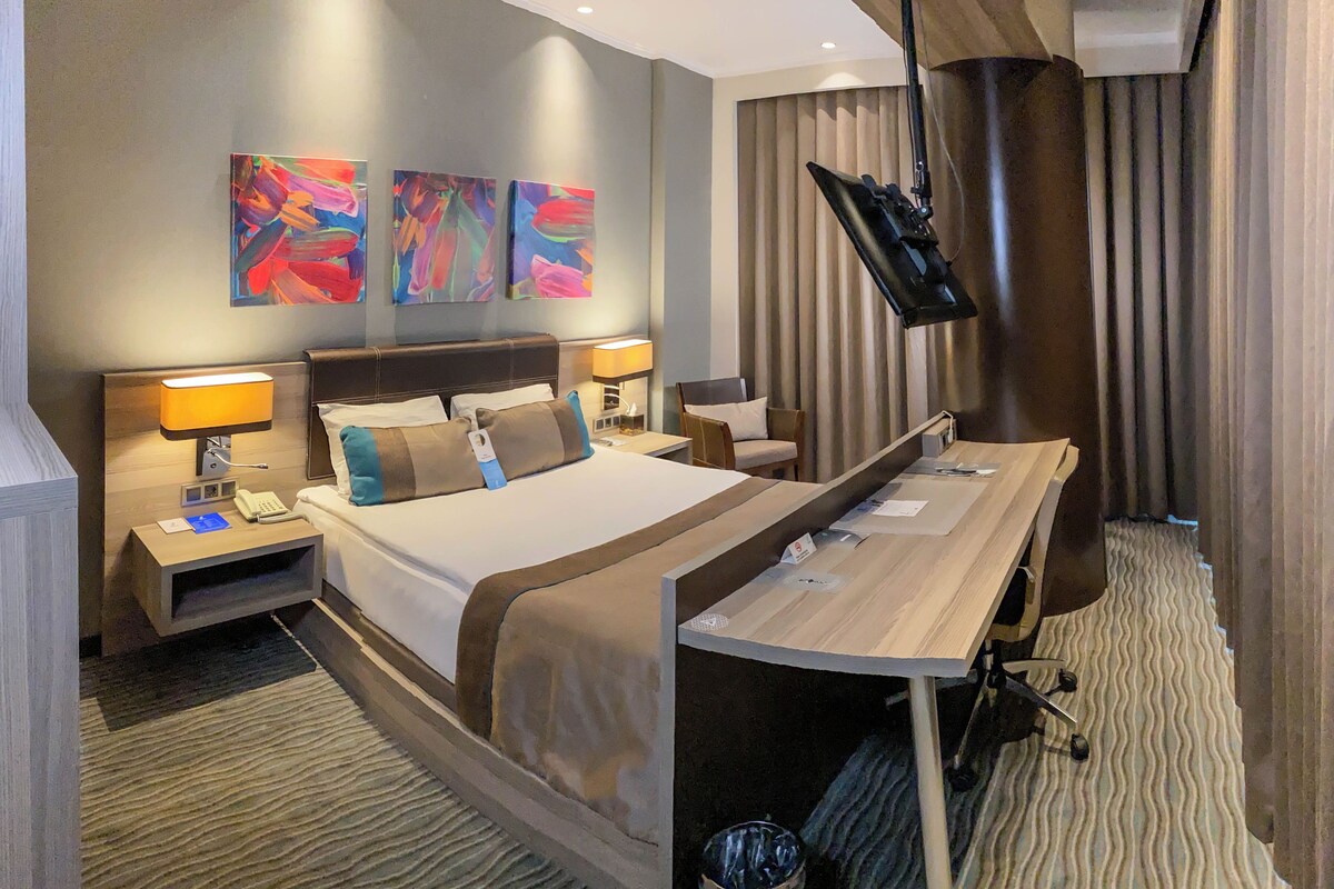 A modern hotel room with a large bed, bedside tables with lamps, a desk, and a flat-screen TV.