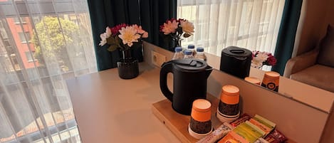 A hotel room with a wooden table, a black coffee maker, a tray with tea bags, and a window with curtains.