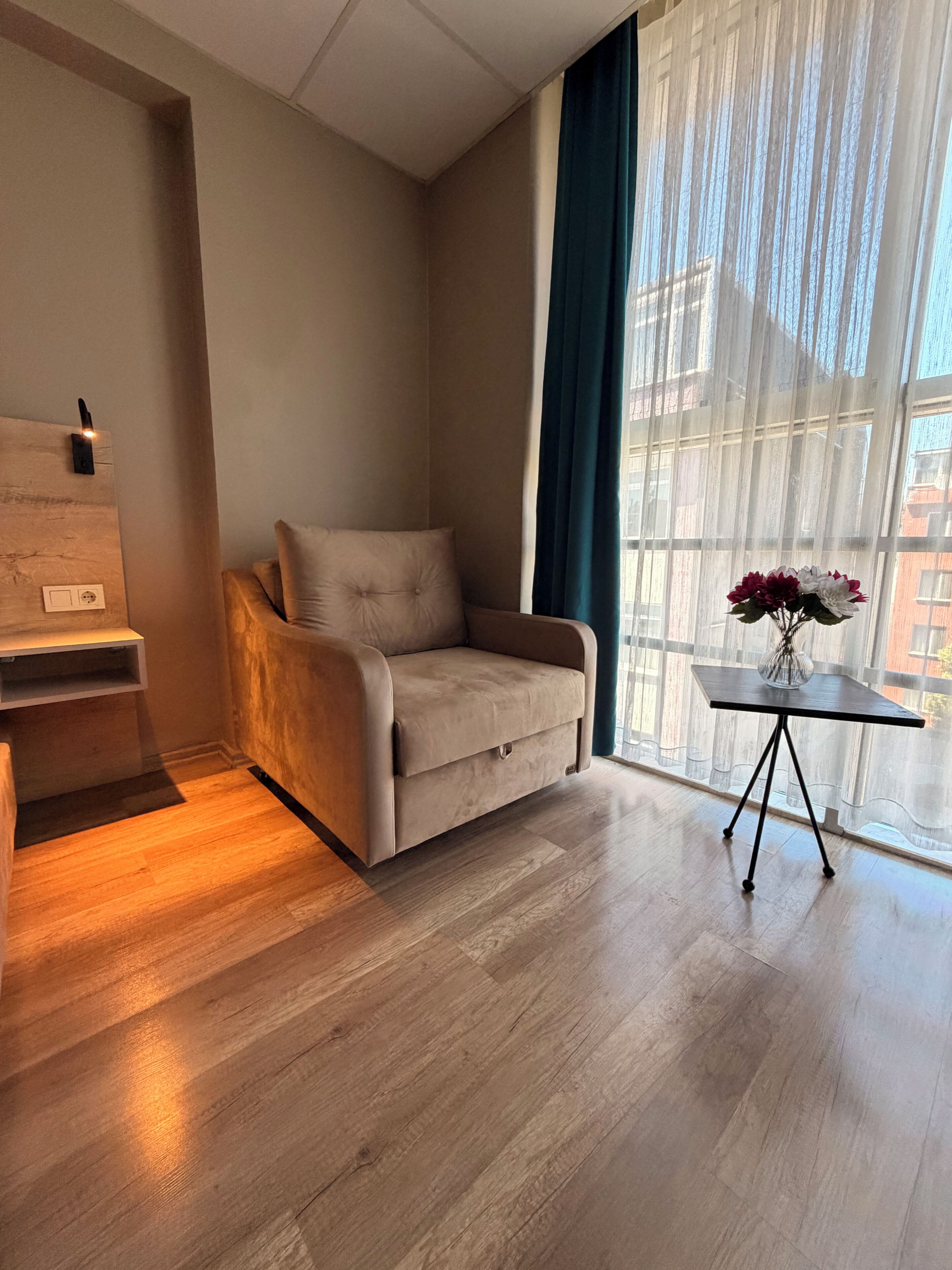 Un salón moderno con un sofá beige, una mesita auxiliar con un jarrón y un ventanal con cortinas traslúcidas.