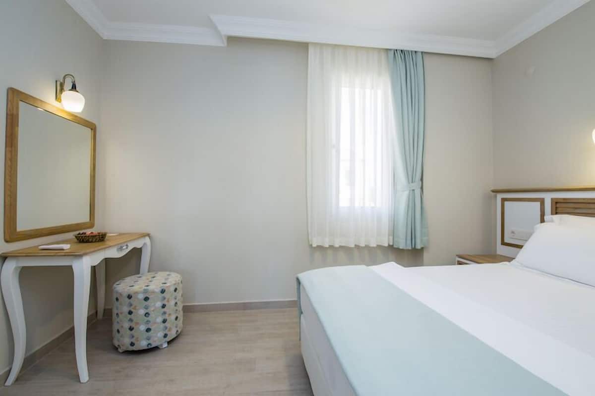 A hotel room with a bed, a bedside table, a mirror, a small stool, and a window with curtains.