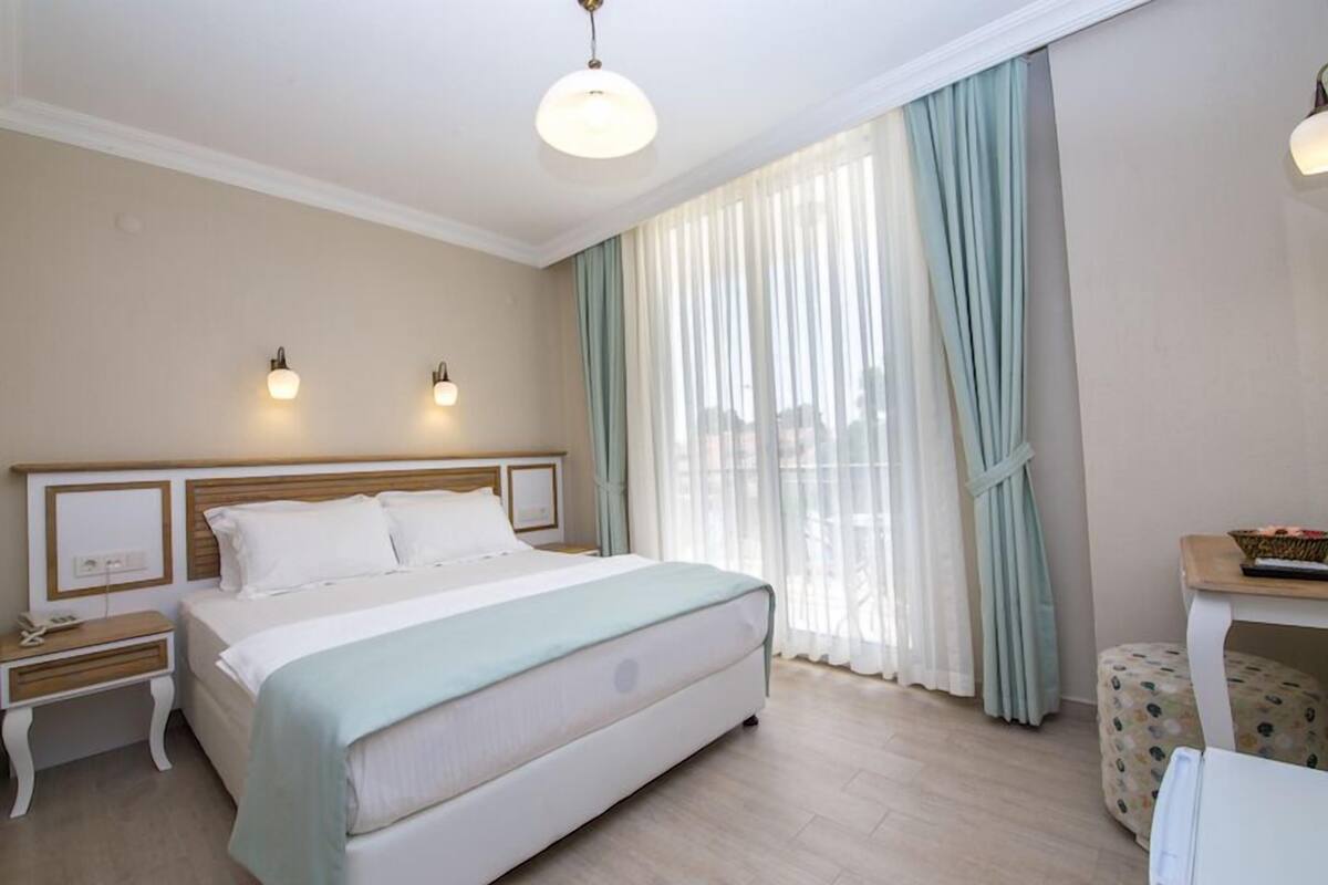 A hotel room with a bed, bedside tables, a nightstand, a lamp, a window with curtains, and a small table with a basket.