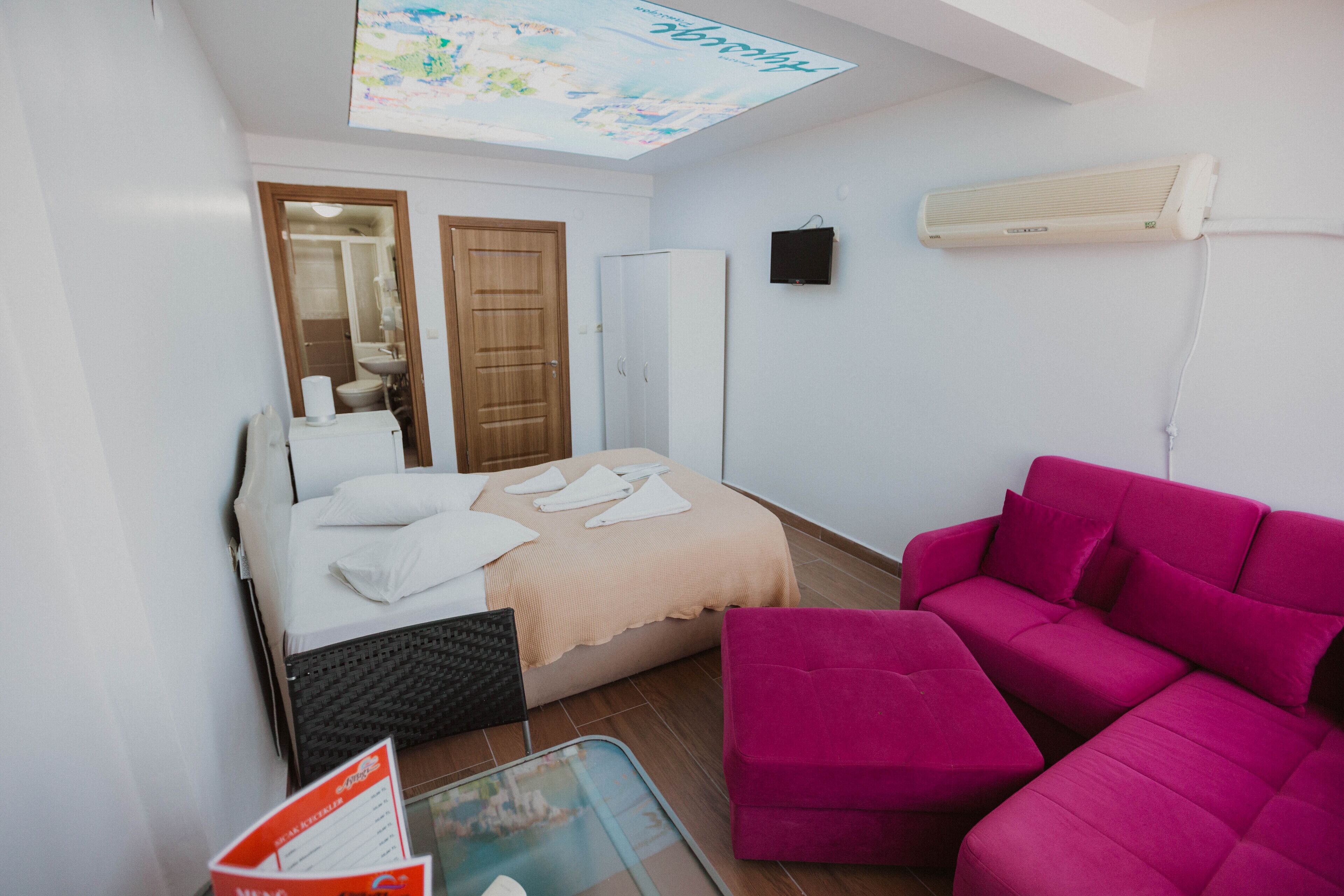 A small, well-lit room with a bed, a pink sofa, a glass table, and a ceiling projector.