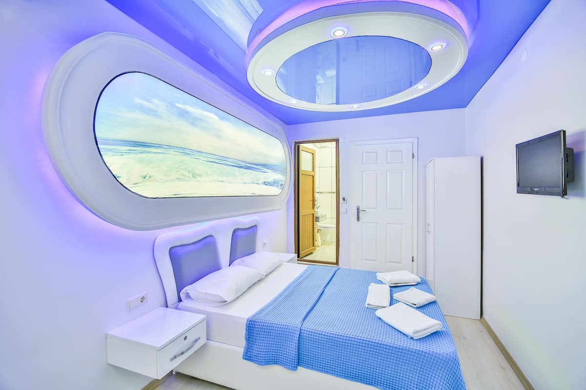 Une chambre moderne avec une grande fenêtre, un lit recouvert d’une couverture bleue, une table de chevet, une armoire et un téléviseur fixé au mur.