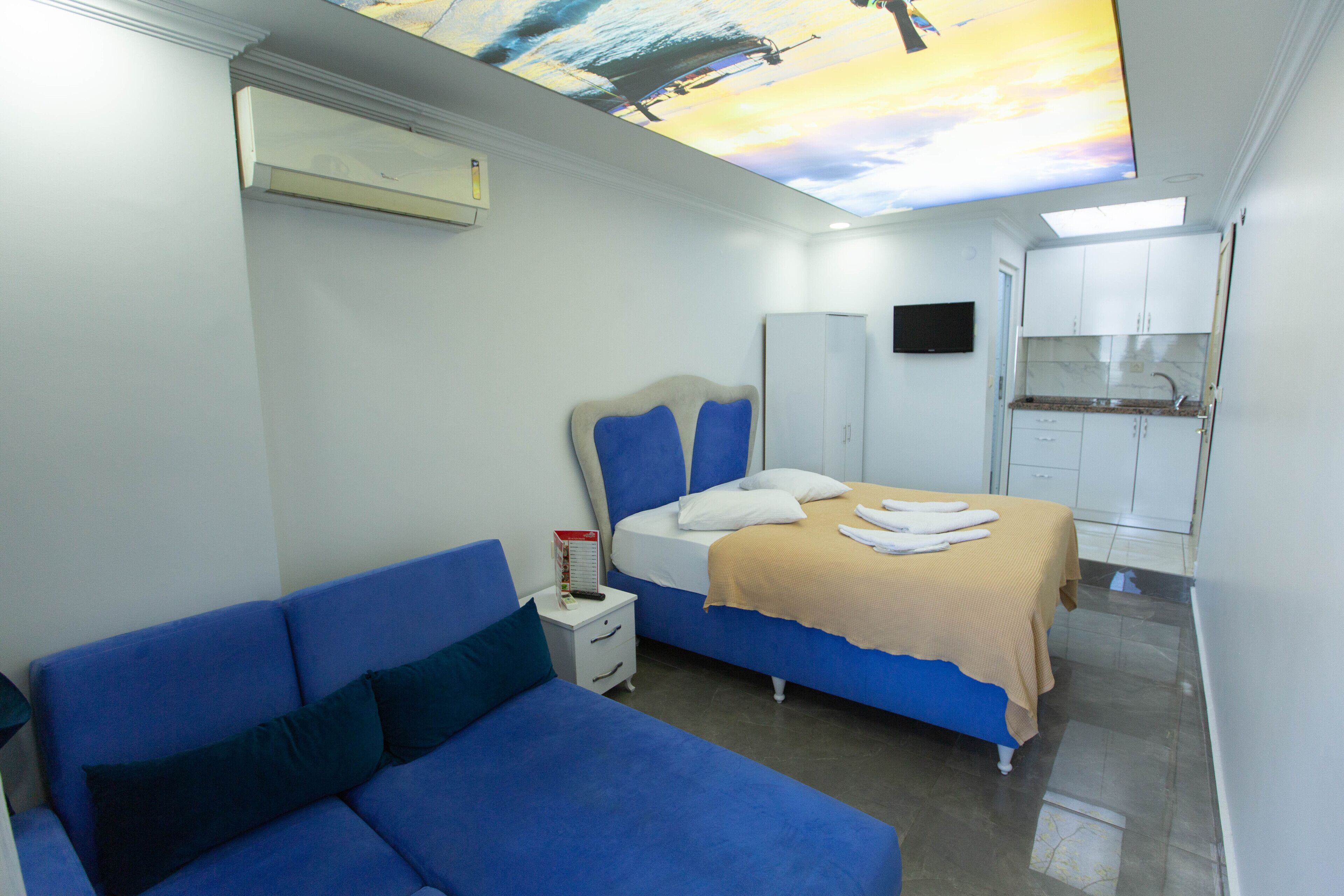 A bedroom with a bed, a sofa, a nightstand, a TV, and a kitchenette.