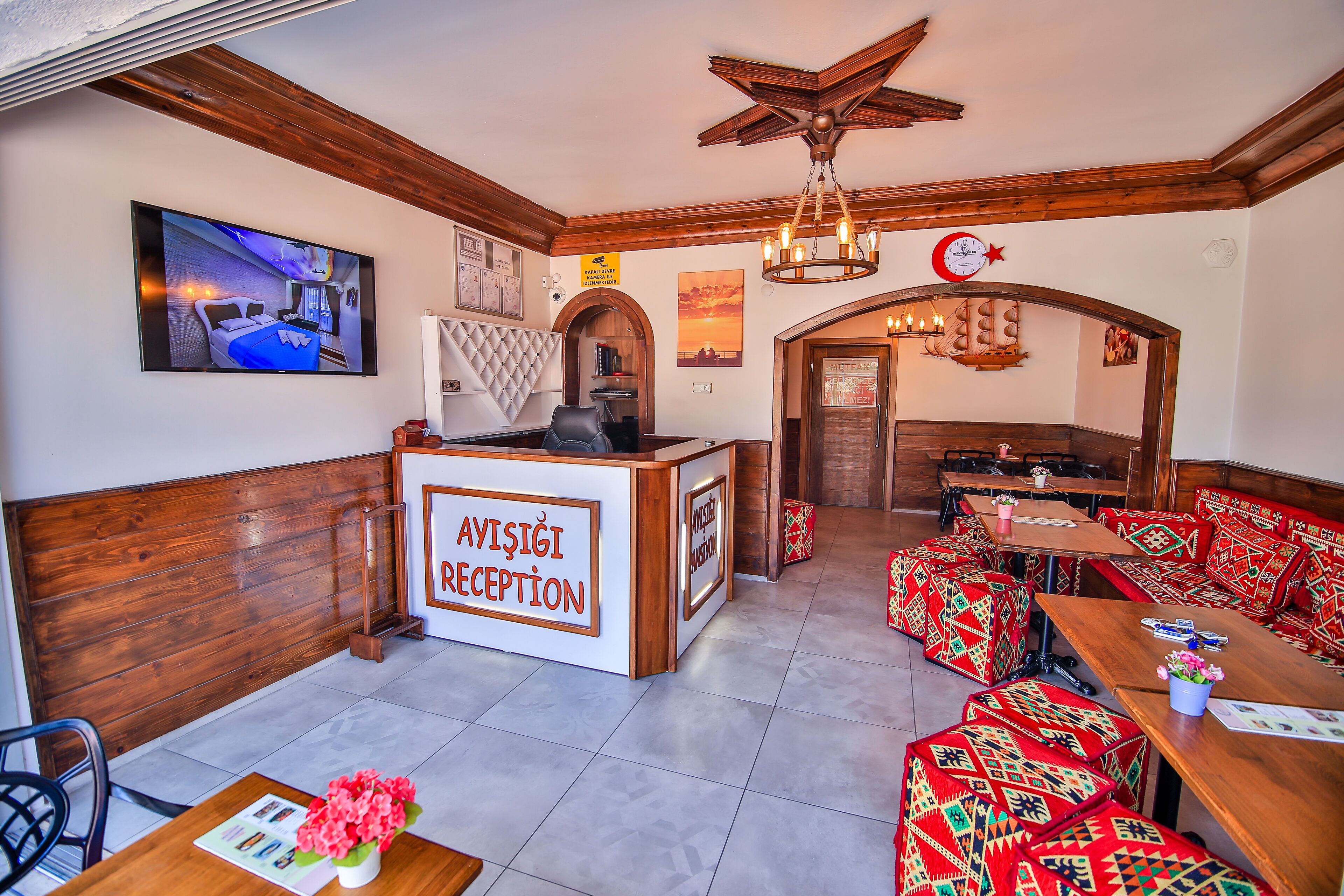 A reception area with a wooden reception desk labeled "AVISIĞI RECEPTION," a television mounted on the wall, and a seating area with patterned cushions.