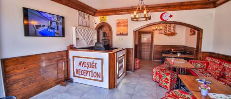 A reception area with a wooden reception desk labeled "AVISIĞI RECEPTION," a television mounted on the wall, and a seating area with patterned cushions.