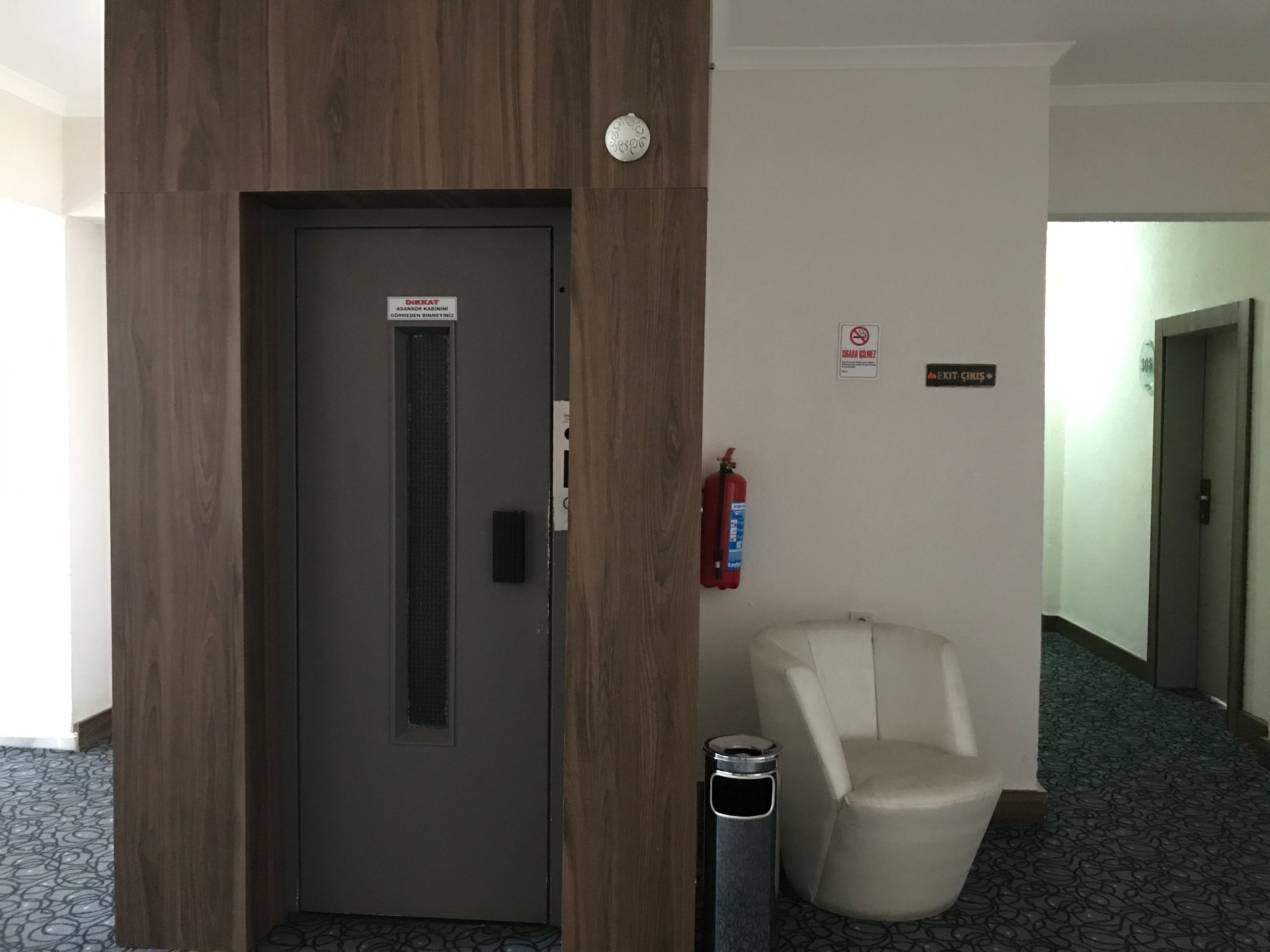Un pasillo de hotel con una puerta de madera, un extintor, una silla blanca y señales indicativas.